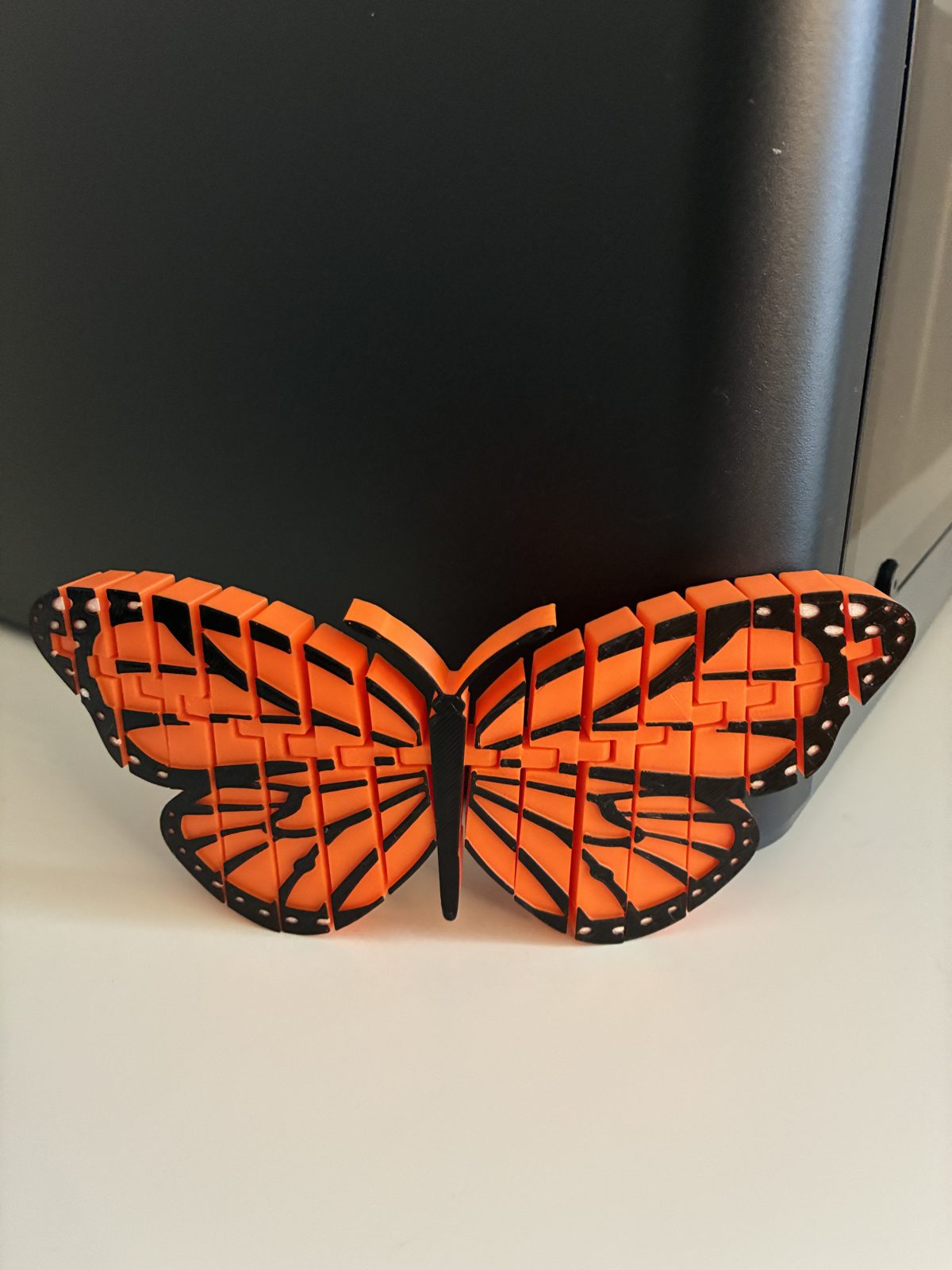 Articulated monarch butterfly printed in orange and black PLA, resting in front of the printer