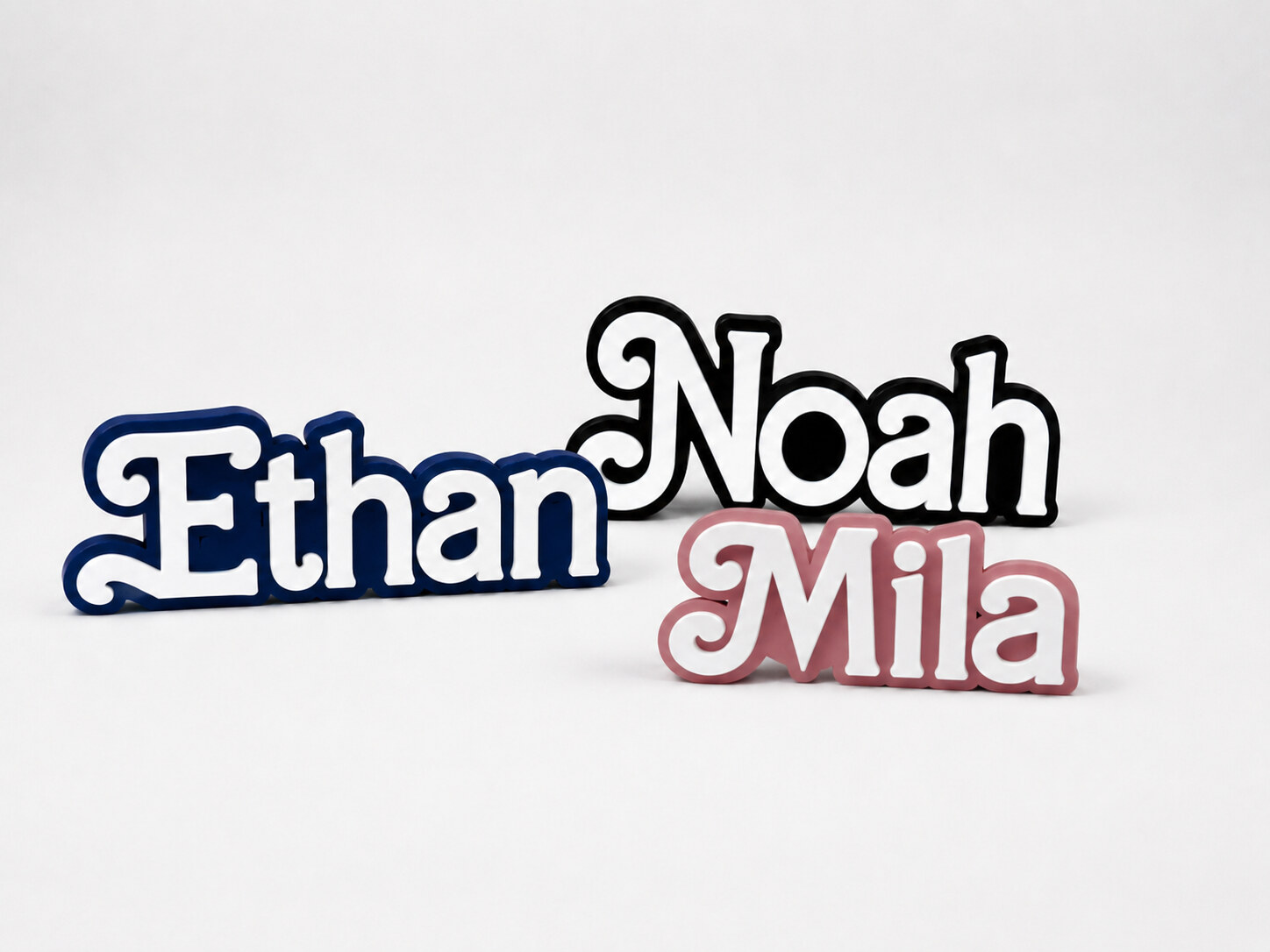 Three custom 3D printed name plates — Ethan in navy blue, Noah in black and Mila in pink, each with a raised white face — arranged on a white backdrop